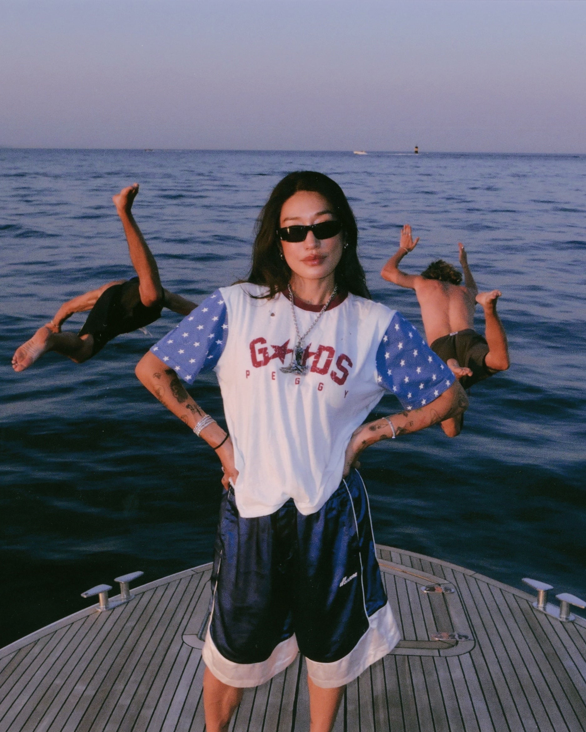 A woman stands on the bow of a boat wearing a white and blue star-patterned t-shirt with 'G*Woods' printed on the front, paired with dark athletic shorts. Two men are seen diving headfirst into the ocean in the background under a soft sunset sky.